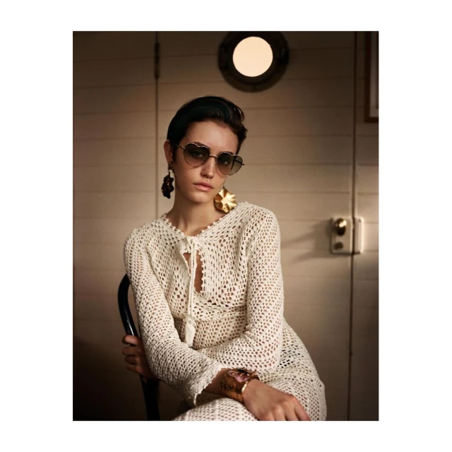 Portrait of a woman wearing a crochet dress, heart-shaped sunglasses, and statement earrings, posing in an indoor setting with soft lighting.