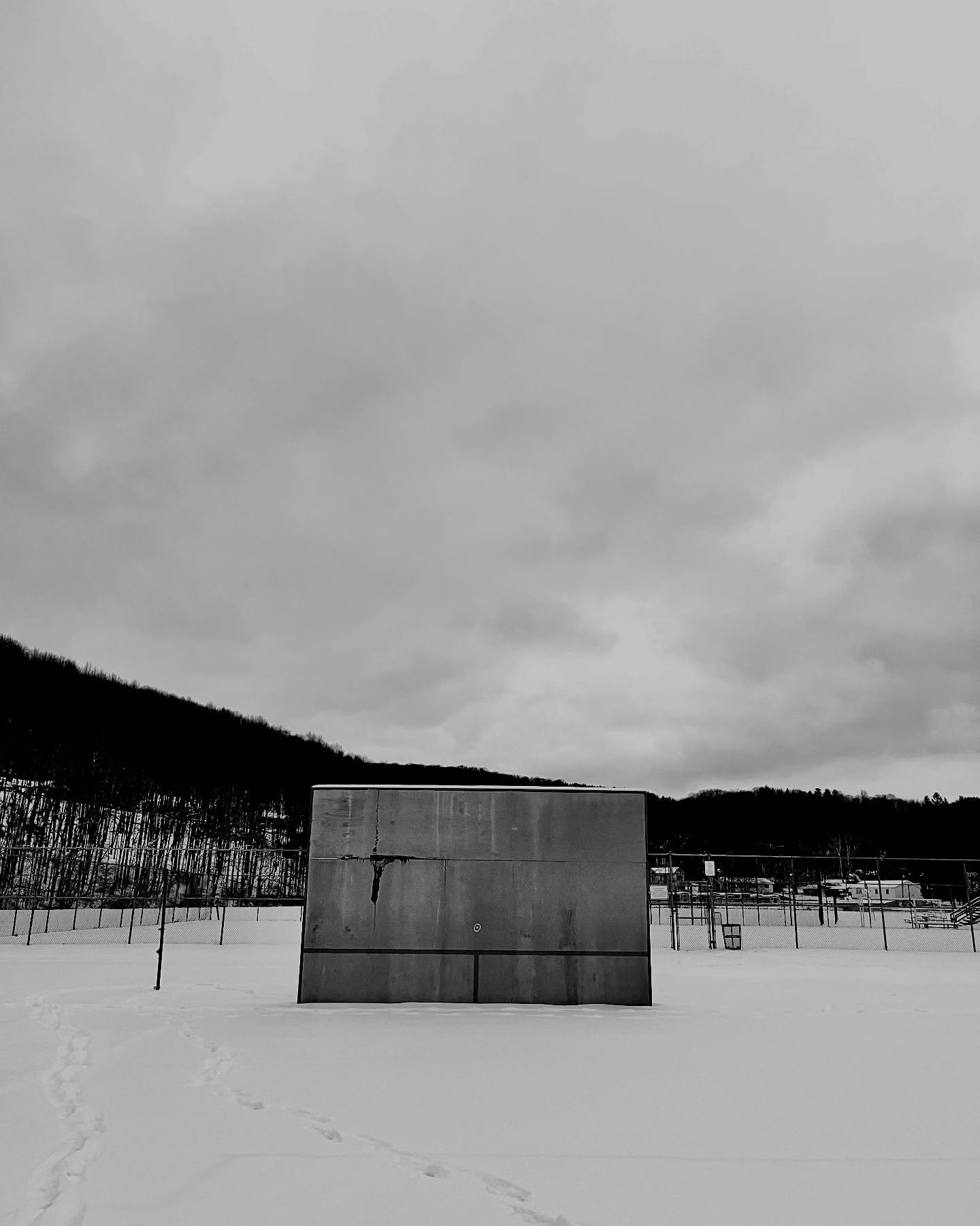 Monochrome photo of a concrete structure in a snow-covered field, set against a backdrop of trees and a cloudy sky.