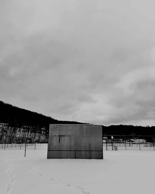Monochrome photo of a concrete structure in a snow-covered field, set against a backdrop of trees and a cloudy sky.