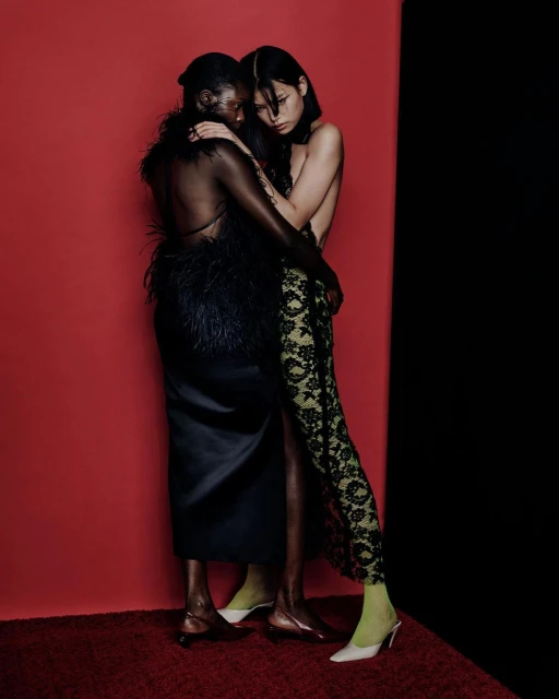 Two women in elegant gowns embrace in a photo studio. One wears a black feathered dress, the other a lace gown and chartreuse mules.