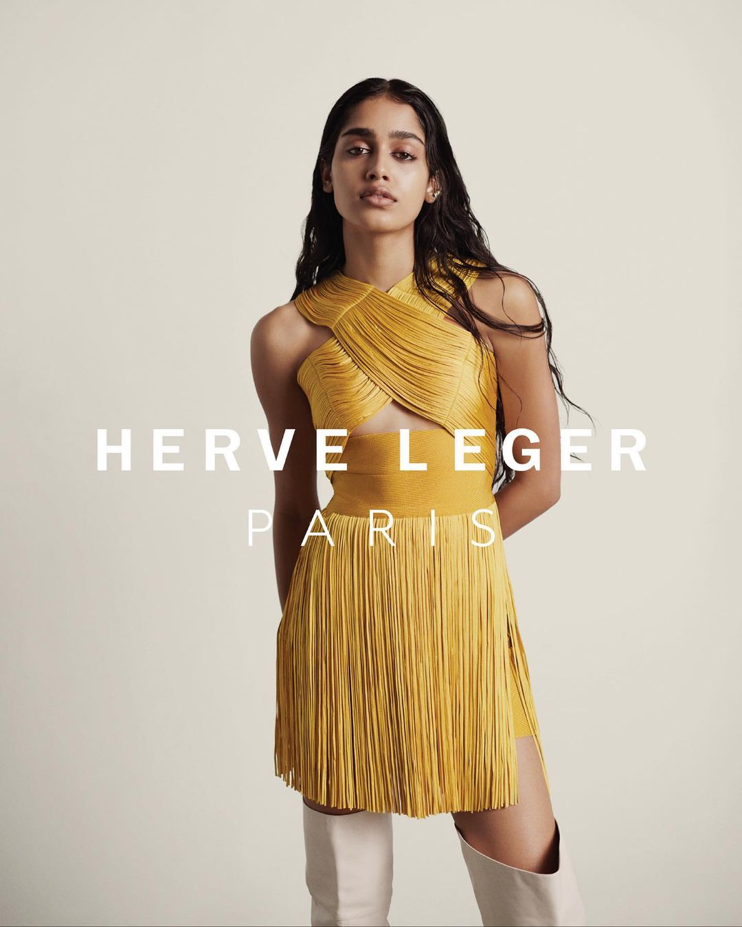 An adult woman poses indoors wearing a yellow fringe dress and white boots, against a cream backdrop with visible brand text.