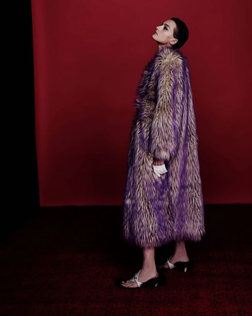 Elegant woman in a luxurious purple and beige fur coat, white gloves, and fashionable sandals posing against a red and black backdrop.