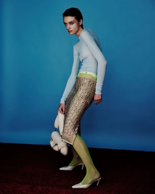 A slender fashion model poses in a light blue top and sequined skirt against a blue backdrop holding a teddy bear purse.