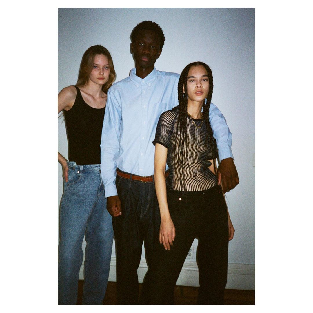 Three fashion models posing indoors in a studio. Two women and one man are standing close to each other. The photograph has neutral light.