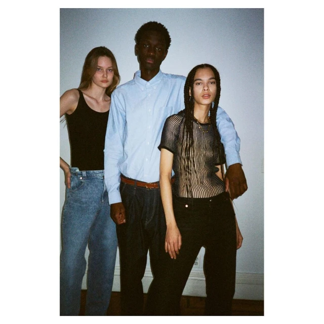 Three fashion models posing indoors in a studio. Two women and one man are standing close to each other. The photograph has neutral light.
