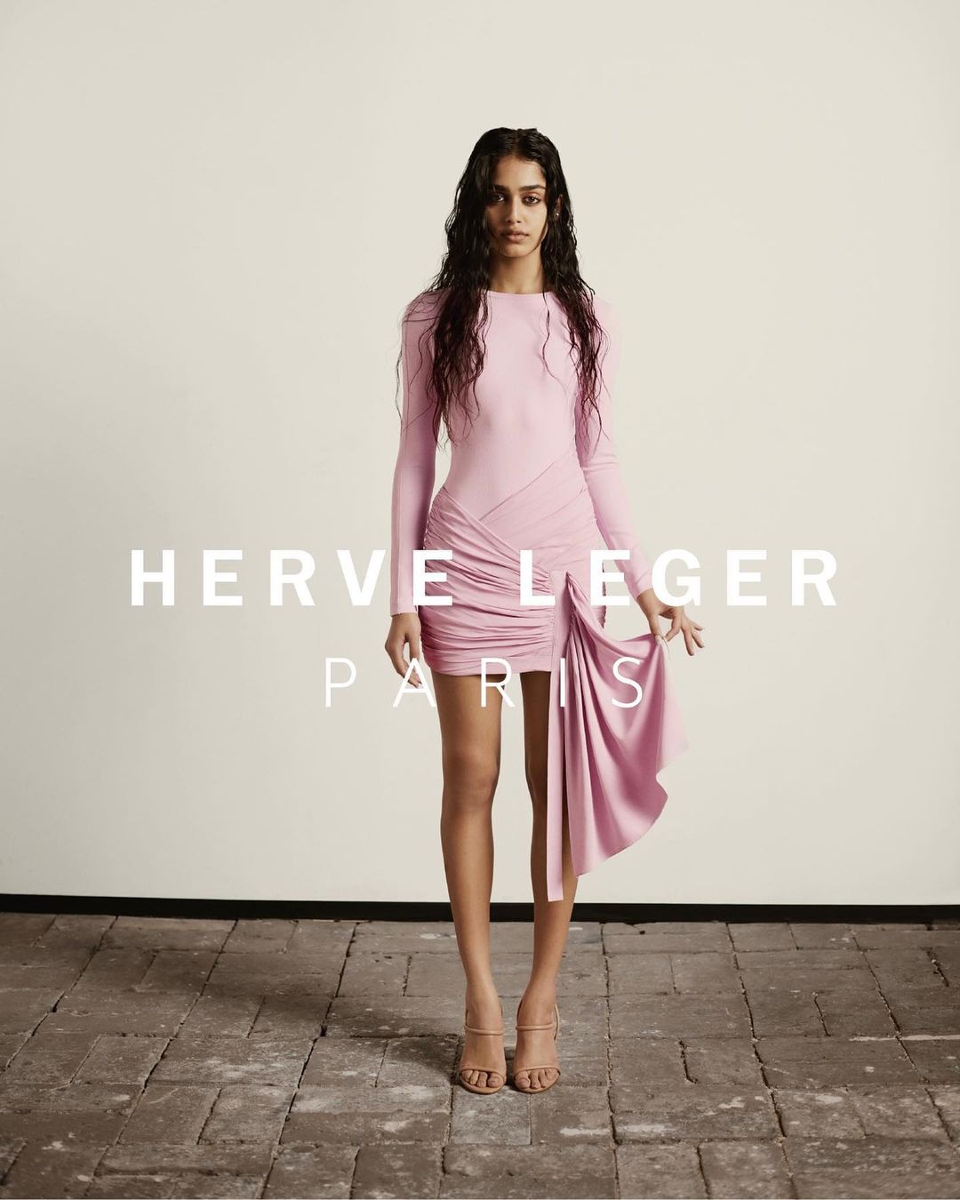Fashionable woman in a light pink Herve Leger mini dress posing in a studio. She wears strappy heels and has long brown hair.
