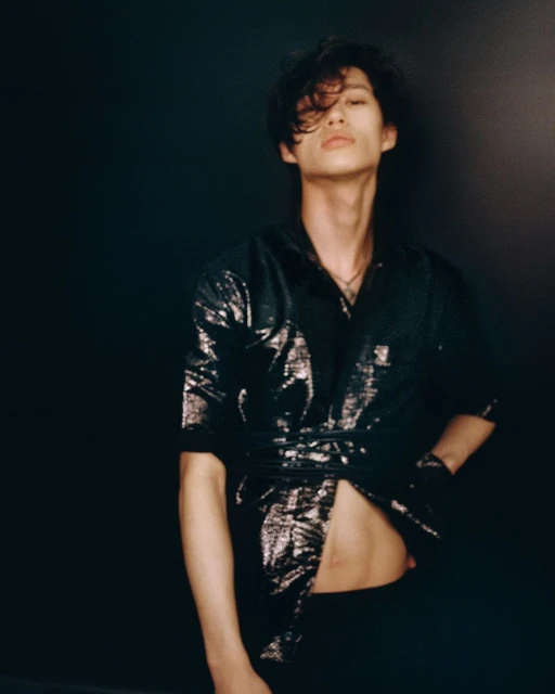 An Asian man with black hair poses against a dark backdrop, wearing a shiny black shirt and pants. He faces forward with an open shirt.