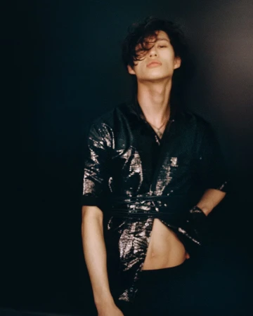 An Asian man with black hair poses against a dark backdrop, wearing a shiny black shirt and pants. He faces forward with an open shirt.