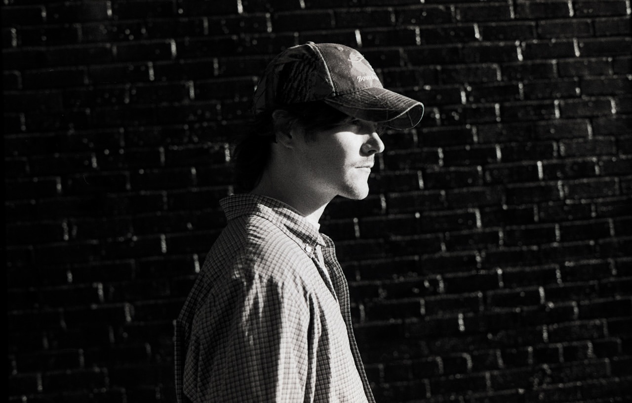 Portrait of an adult male wearing a camouflage hat and button-down shirt, captured in black and white against a brick wall backdrop.