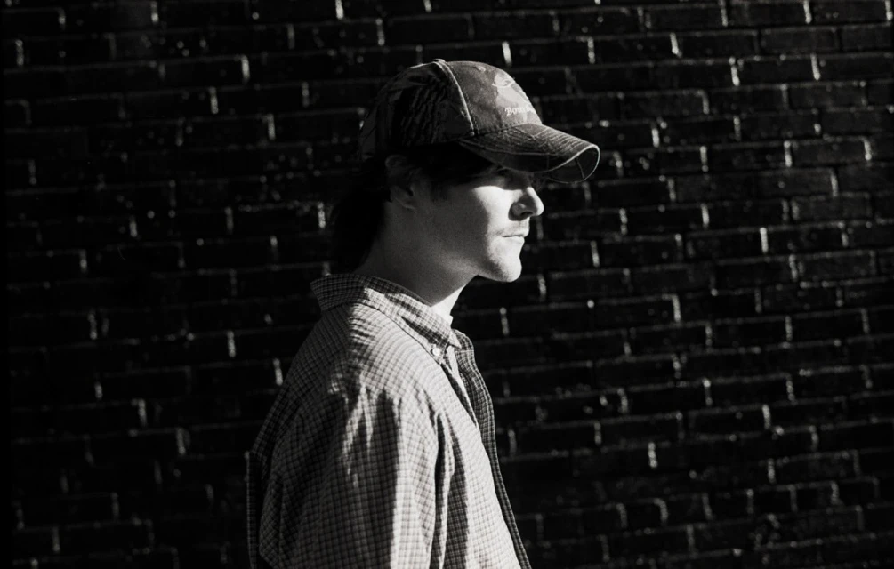 Portrait of an adult male wearing a camouflage hat and button-down shirt, captured in black and white against a brick wall backdrop.