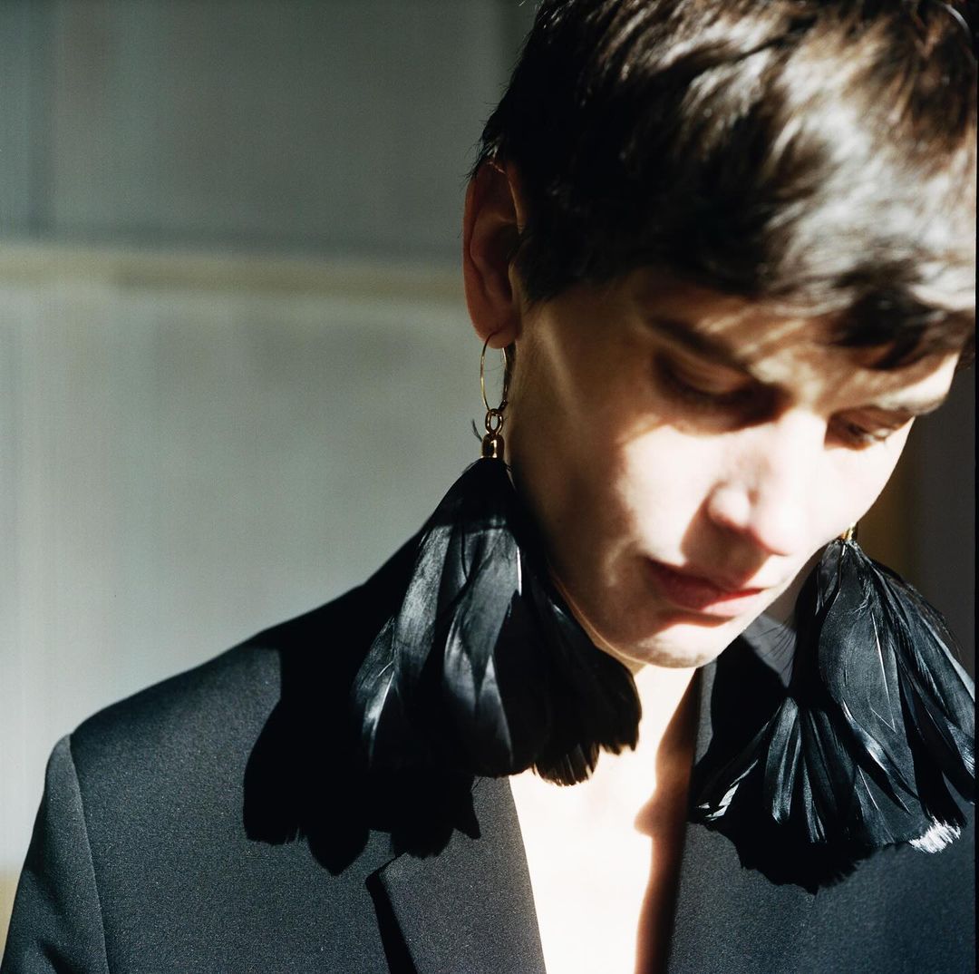 Close-up of a person wearing statement black feather earrings with a gold hoop, black jacket, with natural lighting and minimalist background.