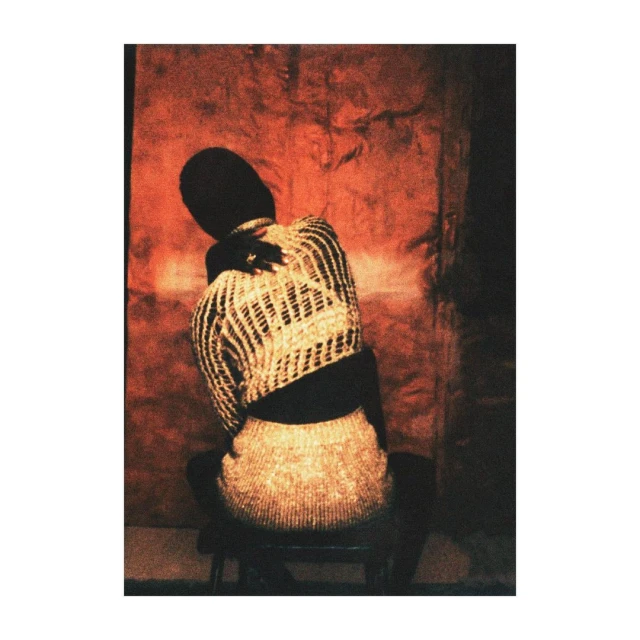 Back view of a Black woman with dark hair, sitting on a stool in a beige and black woven top and skirt with a textured orange background.