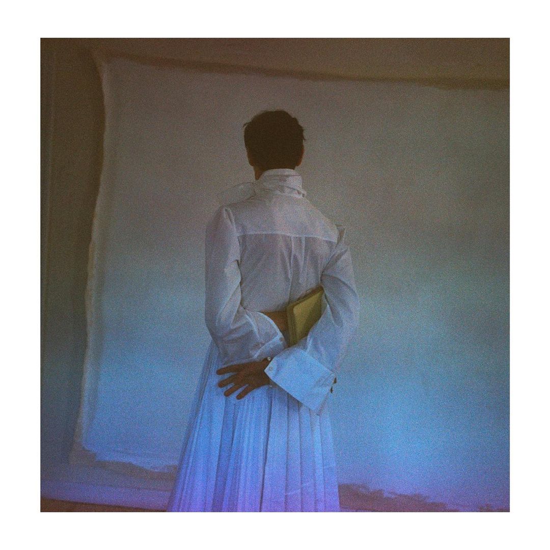 Rear view of a person wearing a white pleated dress and blouse, holding a small gold book against a textured backdrop.