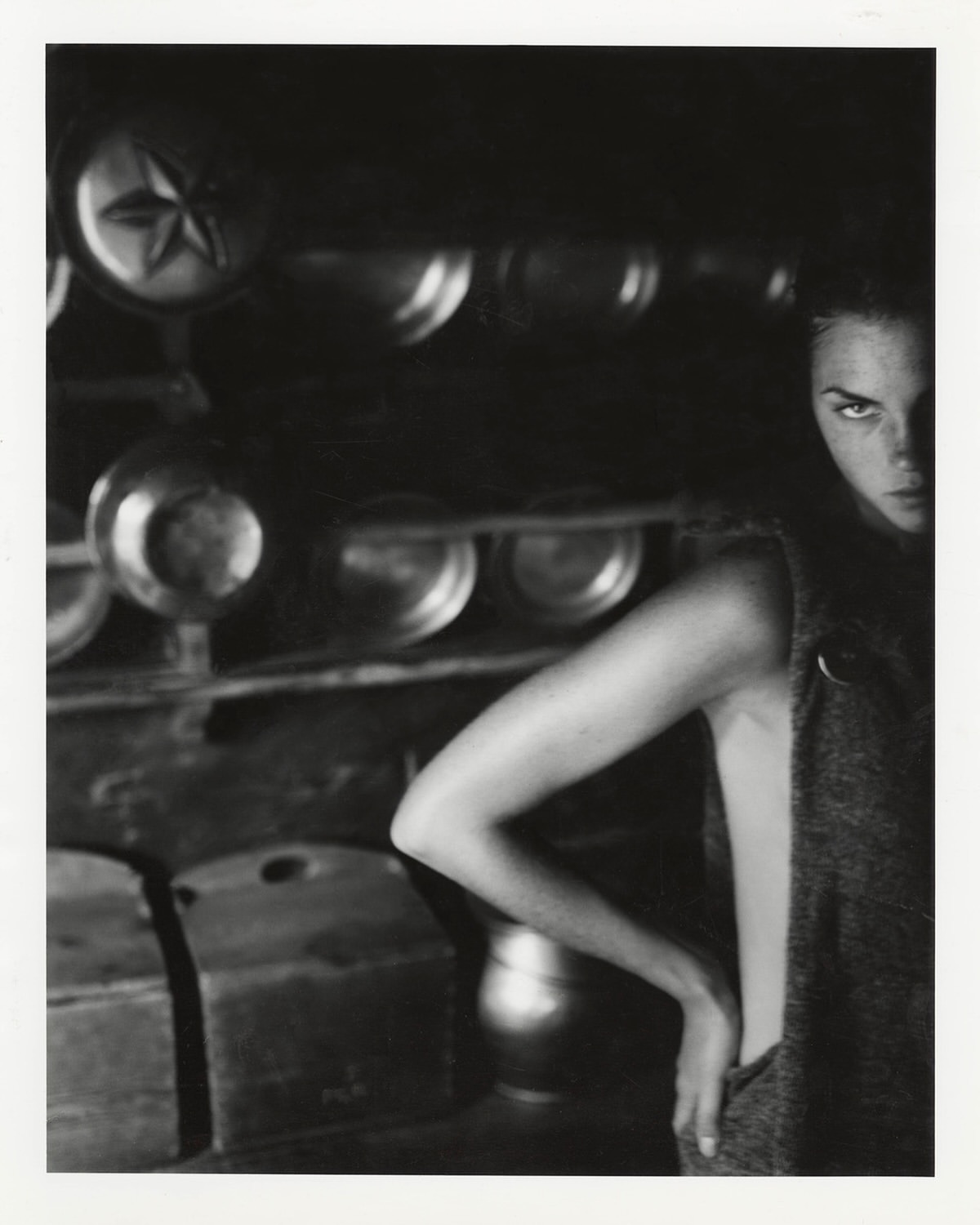 Monochrome portrait of an adult female model in a sleeveless top, gazing intensely towards the camera in a dark, rustic indoor setting.
