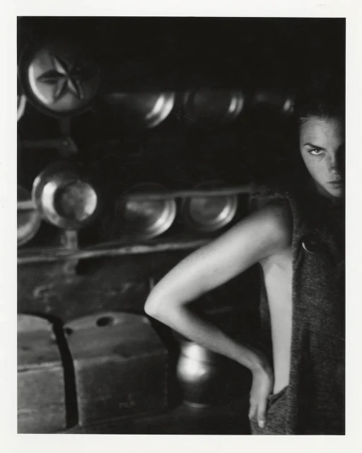 Monochrome portrait of an adult female model in a sleeveless top, gazing intensely towards the camera in a dark, rustic indoor setting.