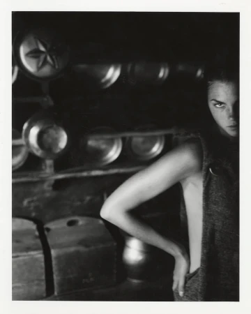 Monochrome portrait of an adult female model in a sleeveless top, gazing intensely towards the camera in a dark, rustic indoor setting.