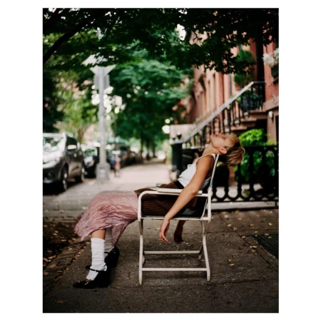Woman in white tank top and pink skirt leaning back in a chair on a city sidewalk by Drew Jarrett