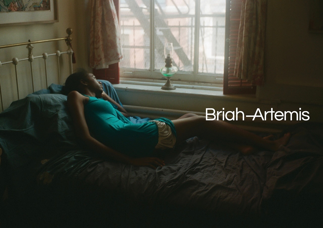 A young woman with dark hair relaxes on a bed in a room with a window, wearing a turquoise dress and tan shorts.