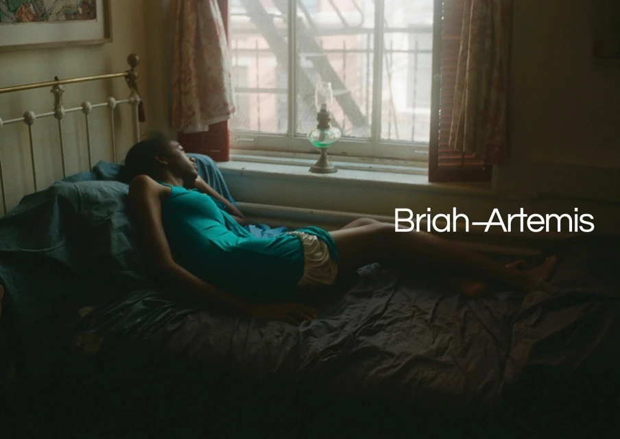 A young woman with dark hair relaxes on a bed in a room with a window, wearing a turquoise dress and tan shorts.
