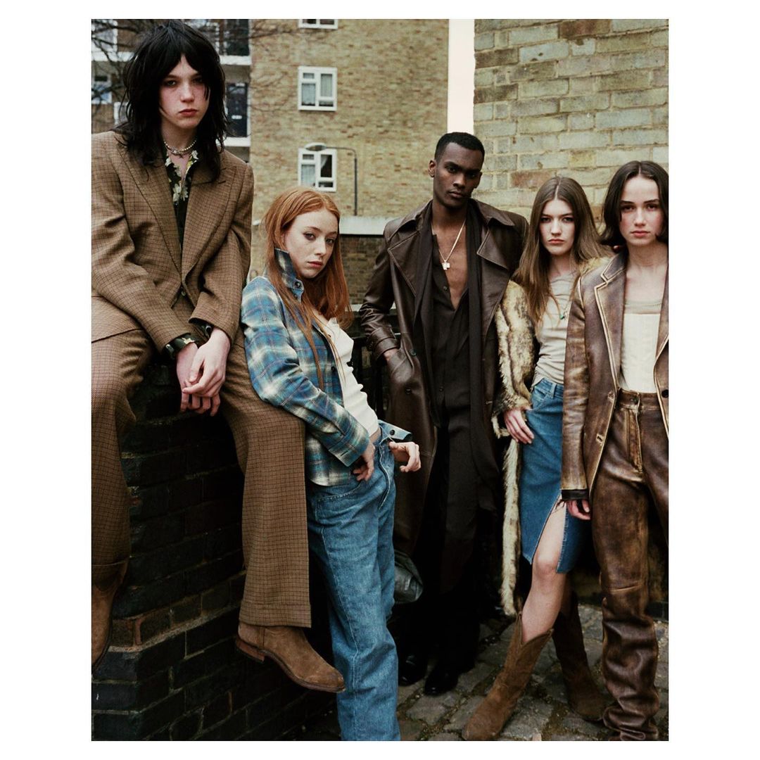 Group fashion shot with five people in vintage-inspired clothing, including a brown coat, blue jeans, and a tan leather jacket outdoors.