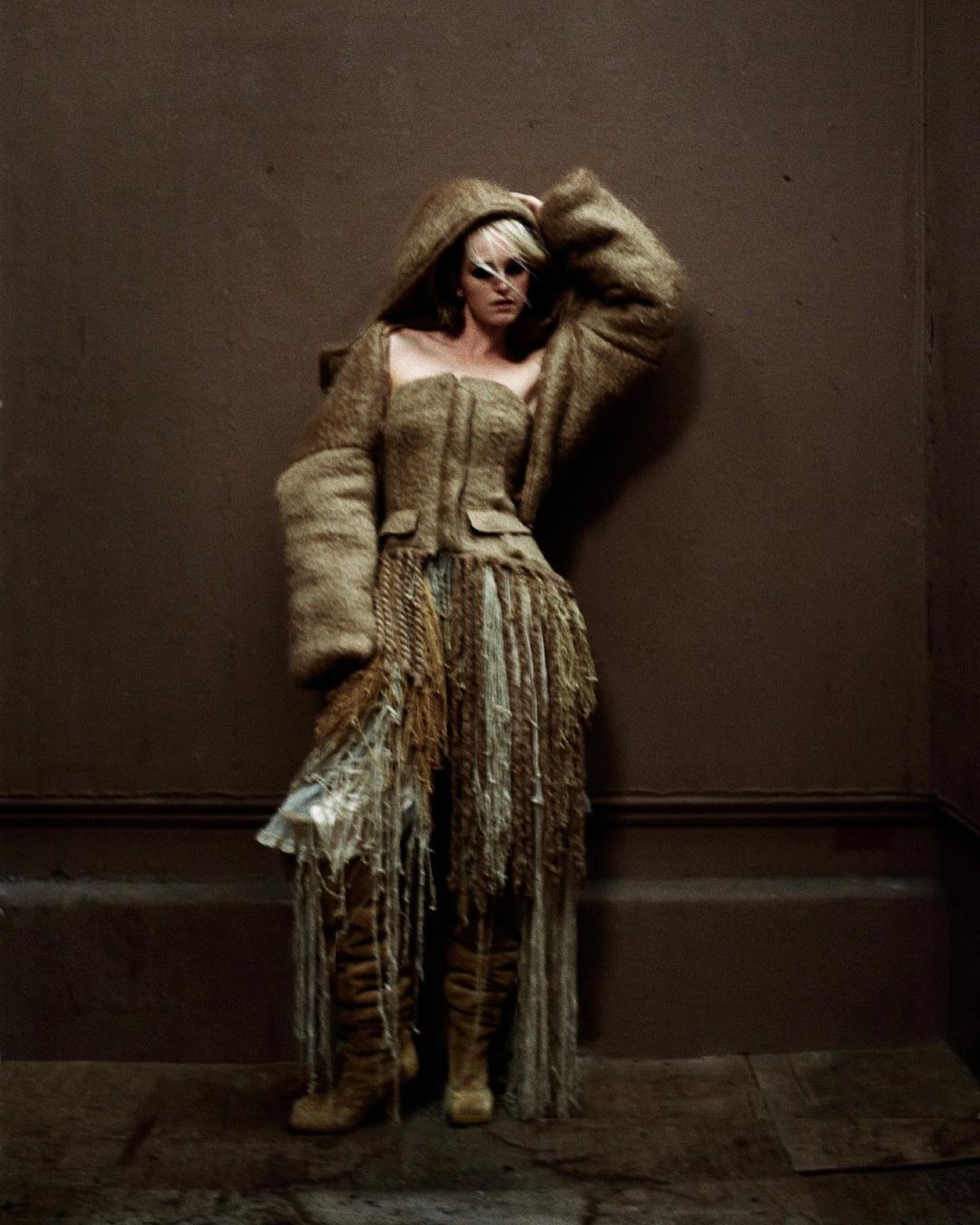 Adult female fashion model poses in a hooded outfit with fringe details against a brown wall and flooring in a studio.