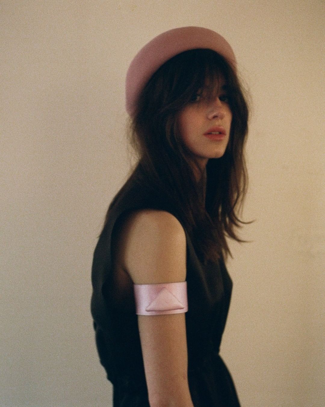 Portrait of a woman with long brown hair wearing a pink hat and arm band against an off-white background in soft studio light.