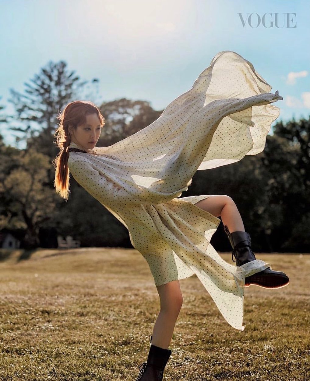 A poised female fashion model in a polka-dot dress and boots, set against a lush, sunlit meadow with distant trees, captured for Vogue.