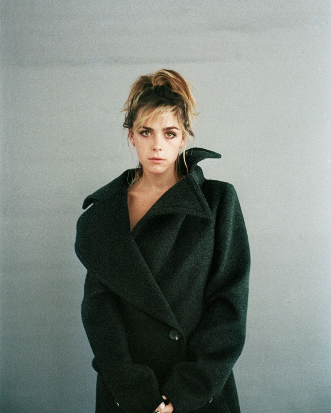 Portrait of a woman with brown hair in a high bun wearing a dark green wool coat against a grey background in an indoor setting.
