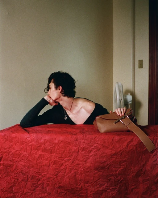 A young adult lies on a red surface next to a tan purse in an indoor setting, with neutral walls and a window fan in the background.