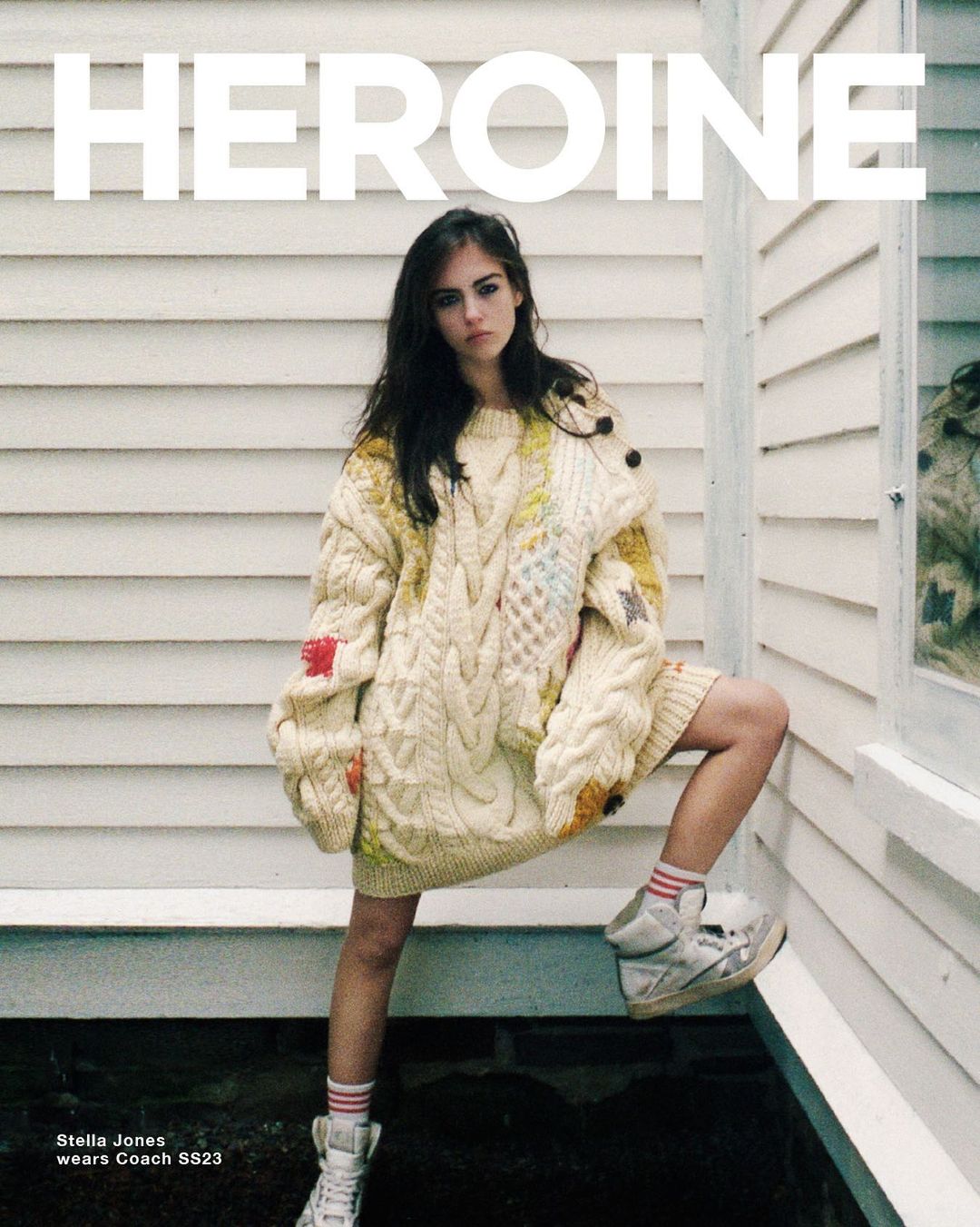 A young woman poses in a chunky, off-white knit sweater and high-top sneakers against a white wood wall, with “HEROINE” text overlay.