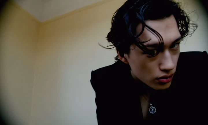 Portrait of a young man with dark wavy hair wearing a black jacket and a silver pendant necklace against a neutral wall.