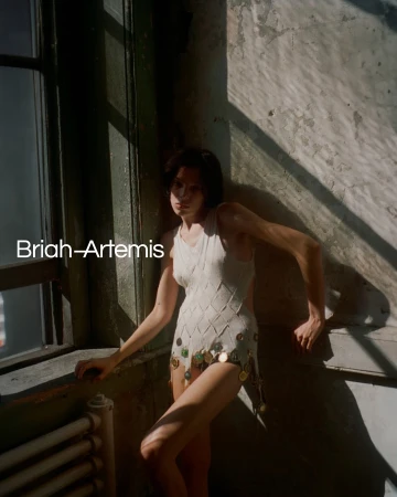 A woman in a white sleeveless patterned top stands near a window against a weathered wall, photographed by Drew Jarrett.