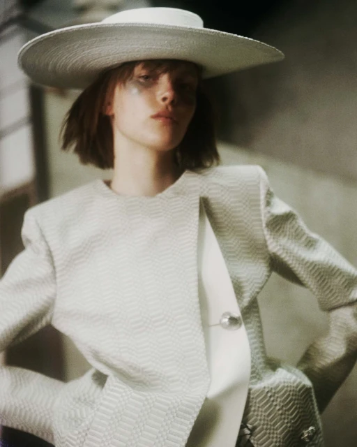 Portrait of a stylish adult woman wearing a wide-brimmed hat and a textured blazer, posing in an indoor environment with soft, diffused lighting.