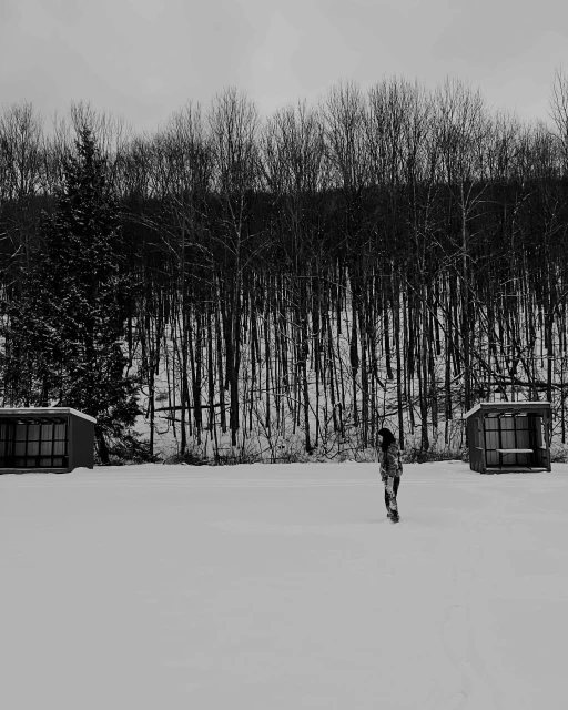 Monochrome photo of a woman walking in a snow-covered landscape near bare trees and small buildings, creating a stark winter scene.