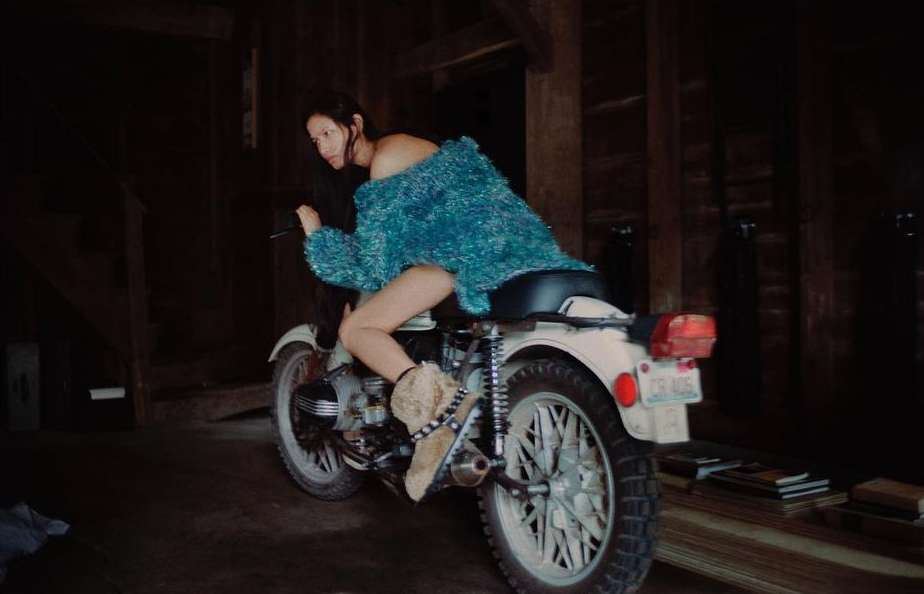 Woman in teal top sits on vintage white motorcycle indoors. Back tire dominates; vintage wooden planks are in the background.