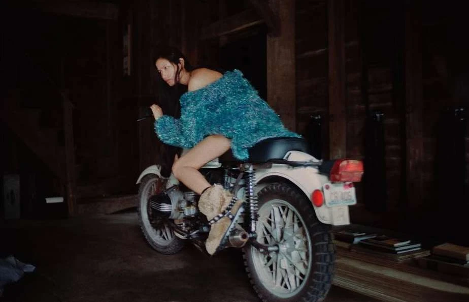 Woman in teal top sits on vintage white motorcycle indoors. Back tire dominates; vintage wooden planks are in the background.