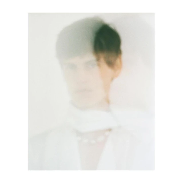 Blurry portrait of a young adult wearing a white top and pearl necklace, set against a soft white background in a diffused light.