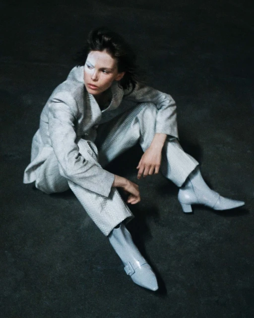 Fashionable woman in grey polka dot suit and light blue boots sitting on grey floor looking left in editorial shot.