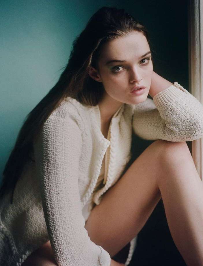 Portrait of a woman wearing a white textured knit cardigan, posed against a dark teal wall with her hand to her head.