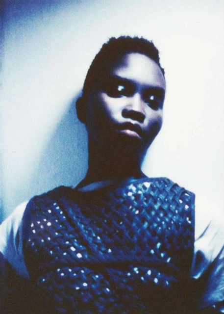 Portrait of a young adult with short hair, wearing a crocheted vest, standing indoors with a neutral facial expression in a blue cast.