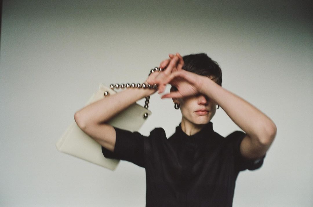 A woman poses indoors with a white purse with silver details, wearing a black top and silver earrings, soft focus, fashion concept.