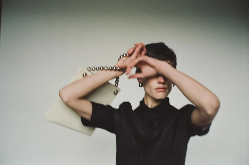 A woman poses indoors with a white purse with silver details, wearing a black top and silver earrings, soft focus, fashion concept.