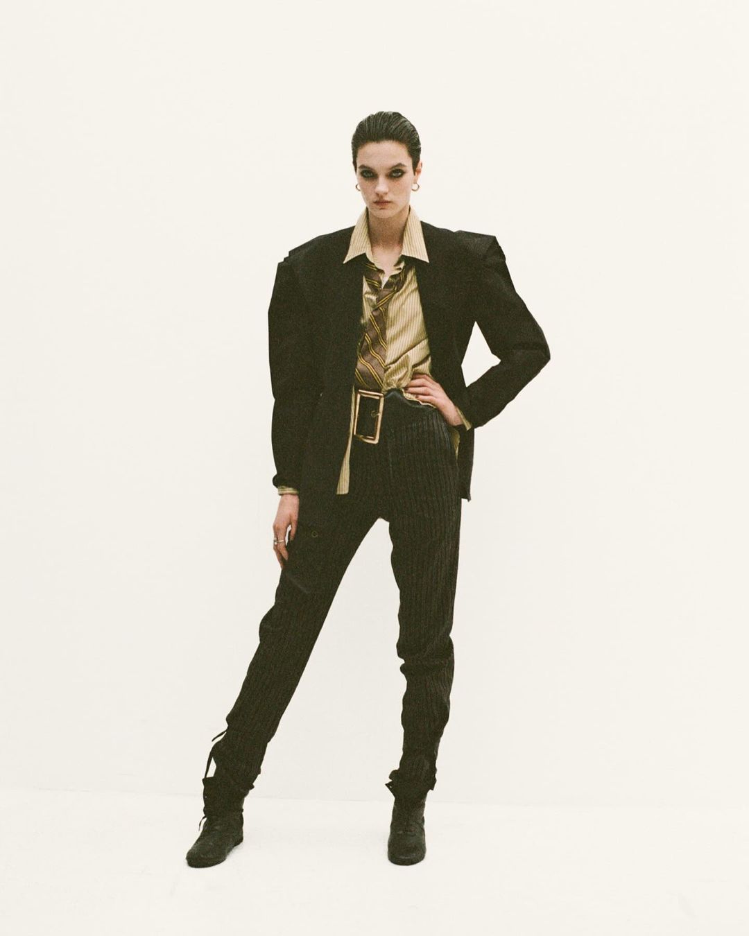 A woman in a tan top, blazer, pinstripe pants, and boots stands in front of a white background in a fashion studio shot.