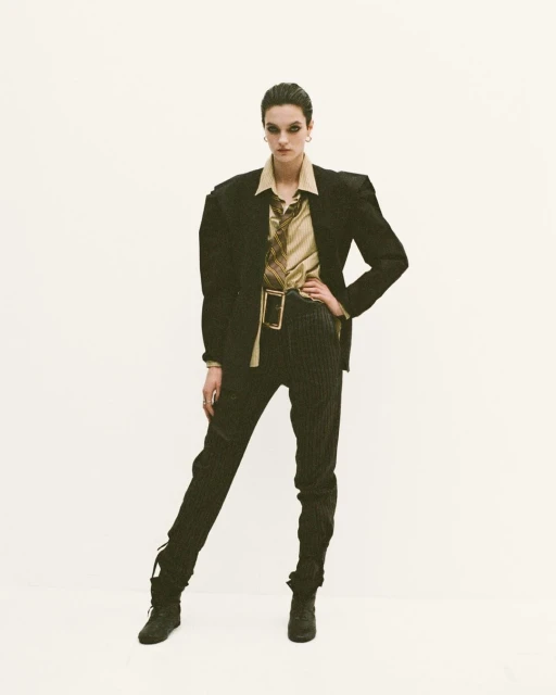 A woman in a tan top, blazer, pinstripe pants, and boots stands in front of a white background in a fashion studio shot.