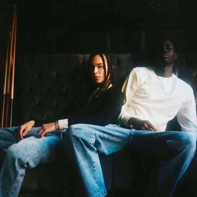 Two people sit on a dark tufted sofa, one with long braids in a black sweater and one in a white shirt and blue jeans.