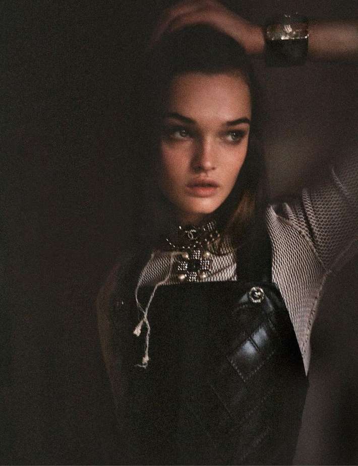 Close-up shot of a woman with dark hair, wearing a leather overall dress and pearl necklace, posing in a dimly lit studio.
