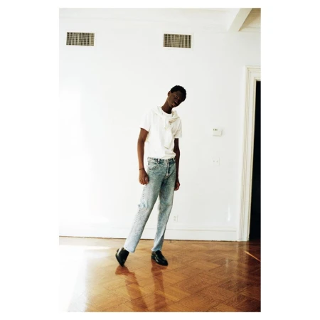 A young adult stands in a relaxed pose in a light-filled room, wearing a white t-shirt, light blue jeans, and black shoes on a parquet floor.