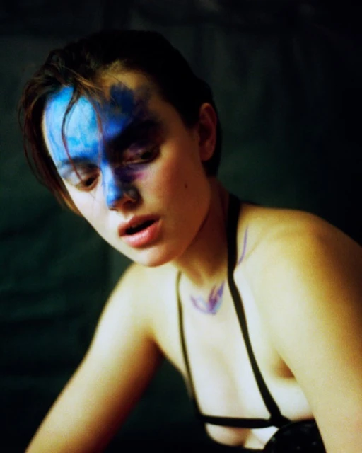 Portrait of a young woman with short hair and blue paint across her face, wearing a black strappy top against a dark background.