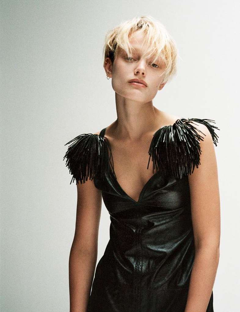 Woman with blond hair wearing a black dress with beaded fringe posing in a studio with a white background for a fashion editorial.