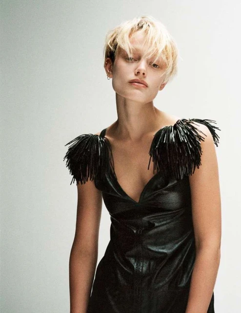 Woman with blond hair wearing a black dress with beaded fringe posing in a studio with a white background for a fashion editorial.